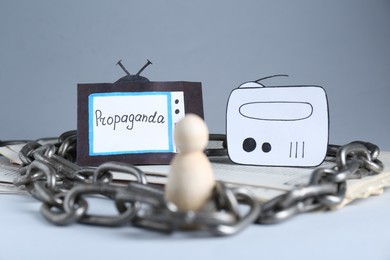 Propaganda concept. Human mired in media field. Chained wooden figure, newspapers, paper TV and radio on table