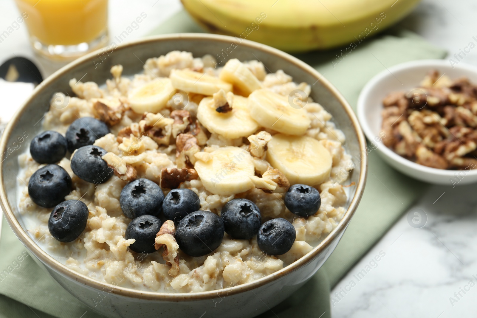 Photo of Tasty oatmeal with banana, blueberries, walnuts and milk served in bowl on white marble table, closeup