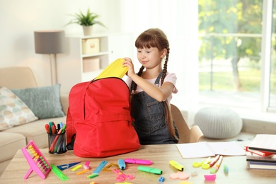 Photo of Cute girl putting school stationery into backpack at table indoors