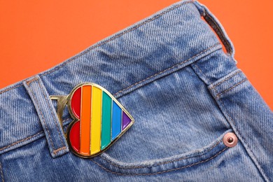 Jeans with rainbow heart shaped pendant on orange background, top view. LGBT pride
