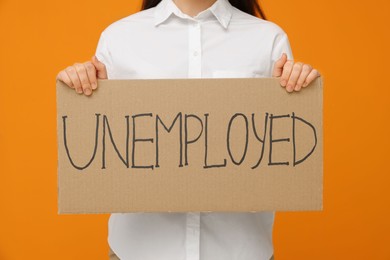 Photo of Young woman holding sign with word Unemployed on orange background, closeup