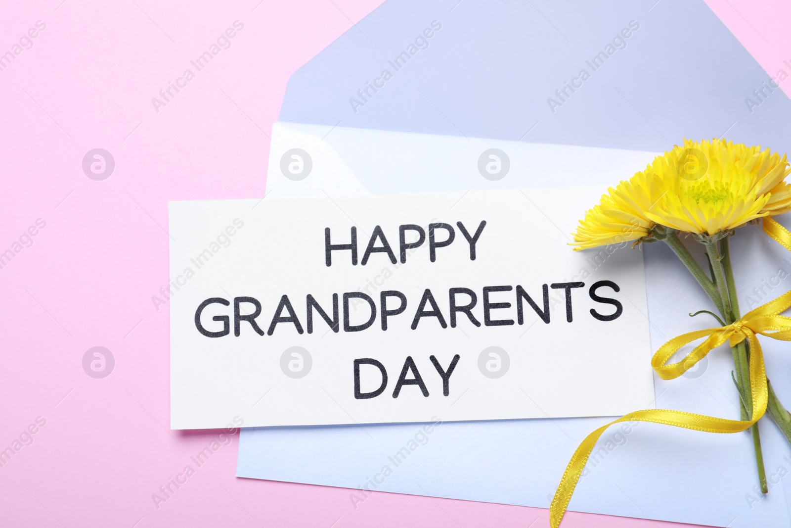 Photo of Card with phrase Happy Grandparents Day, envelope and beautiful flowers on pink background, flat lay