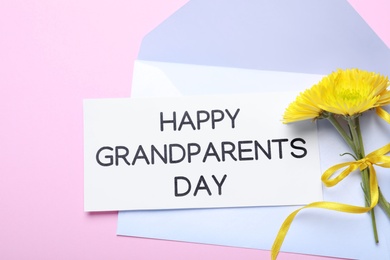 Photo of Card with phrase Happy Grandparents Day, envelope and beautiful flowers on pink background, flat lay