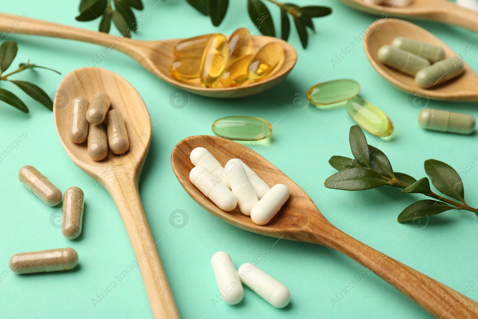 Photo of Different vitamin capsules in spoons and leaves on turquoise background, closeup