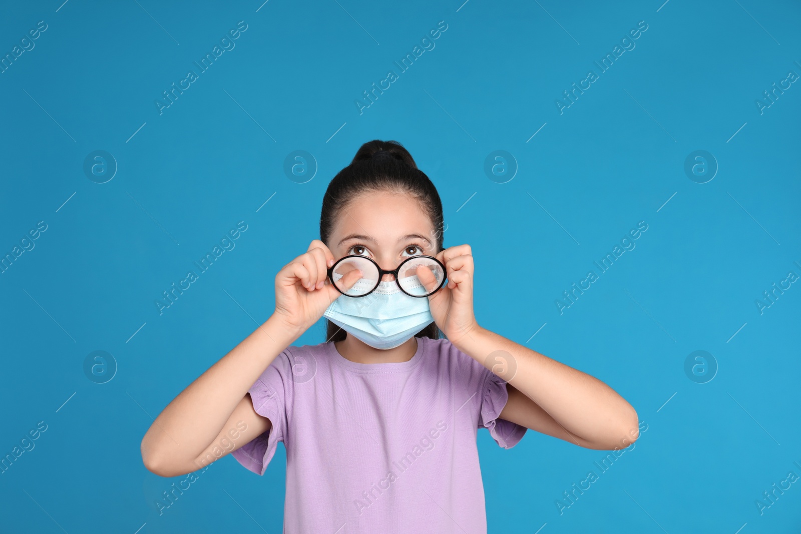 Photo of Little girl wiping foggy glasses caused by wearing medical face mask on blue background. Protective measure during coronavirus pandemic