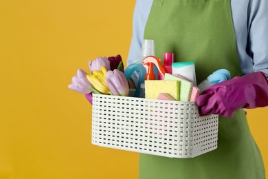 Photo of Spring cleaning. Woman holding basket with detergents, flowers and tools on orange background, closeup. Space for text