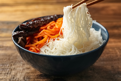 Chopsticks with tasty cooked rice noodles over bowl on wooden table, closeup