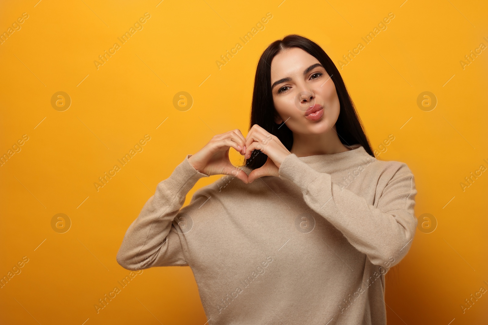 Photo of Beautiful young woman making heart with hands and blowing kiss on orange background. Space for text