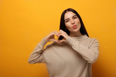 Photo of Beautiful young woman making heart with hands and blowing kiss on orange background. Space for text