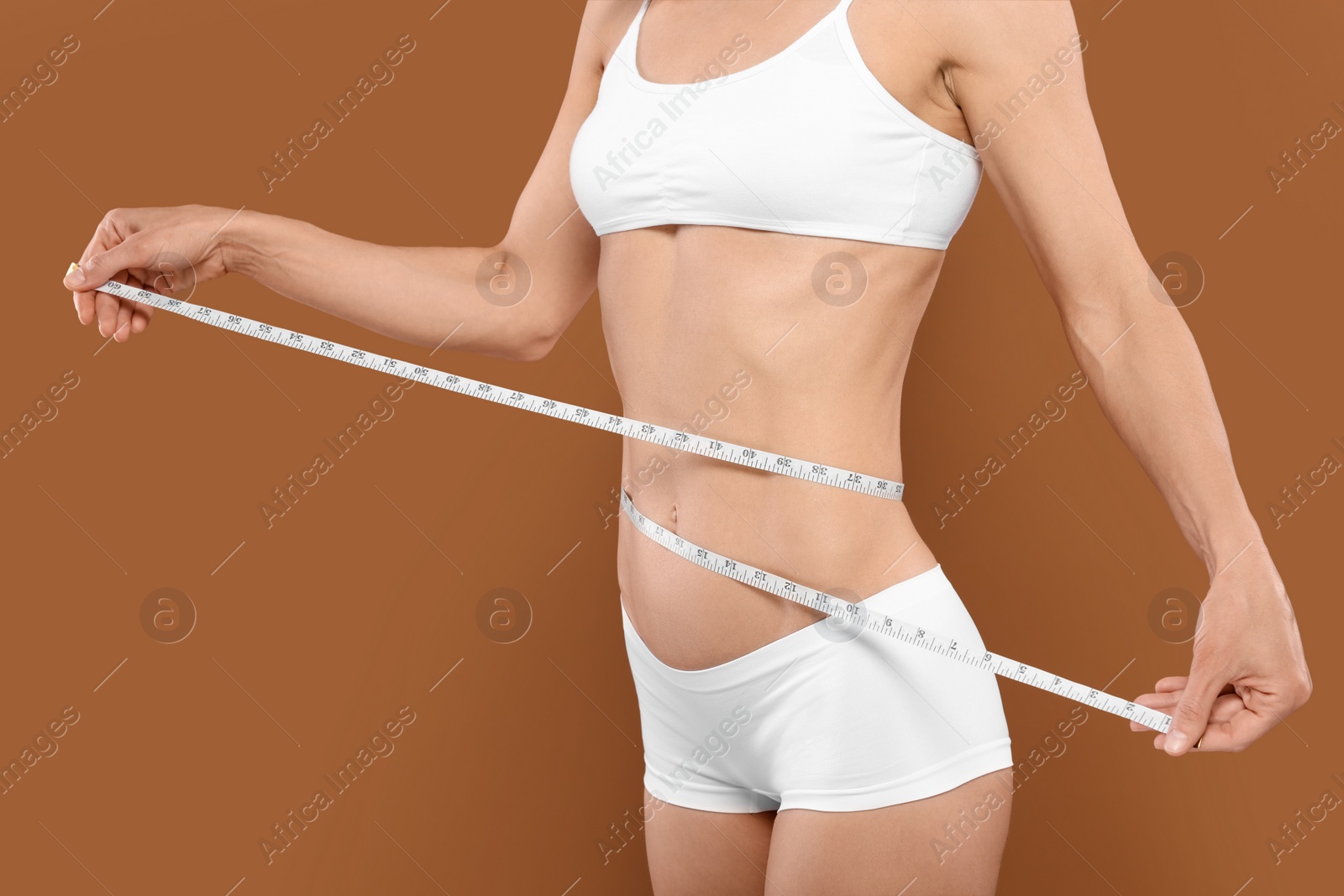 Photo of Slim woman measuring waist with tape on brown background, closeup. Weight loss