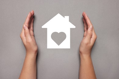 Image of Woman holding hands around paper silhouette of house on grey background, top view. Insurance concept