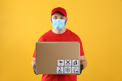 Photo of Courier in mask holding cardboard box with different packaging symbols on yellow background. Parcel delivery