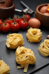 Photo of Uncooked tagliatelle and fresh ingredients on black table, closeup
