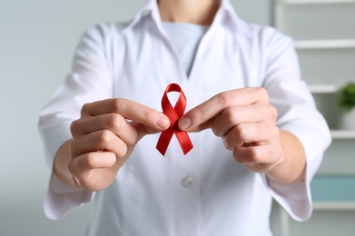 Photo of Doctor holding red awareness ribbon indoors, closeup. World AIDS disease day