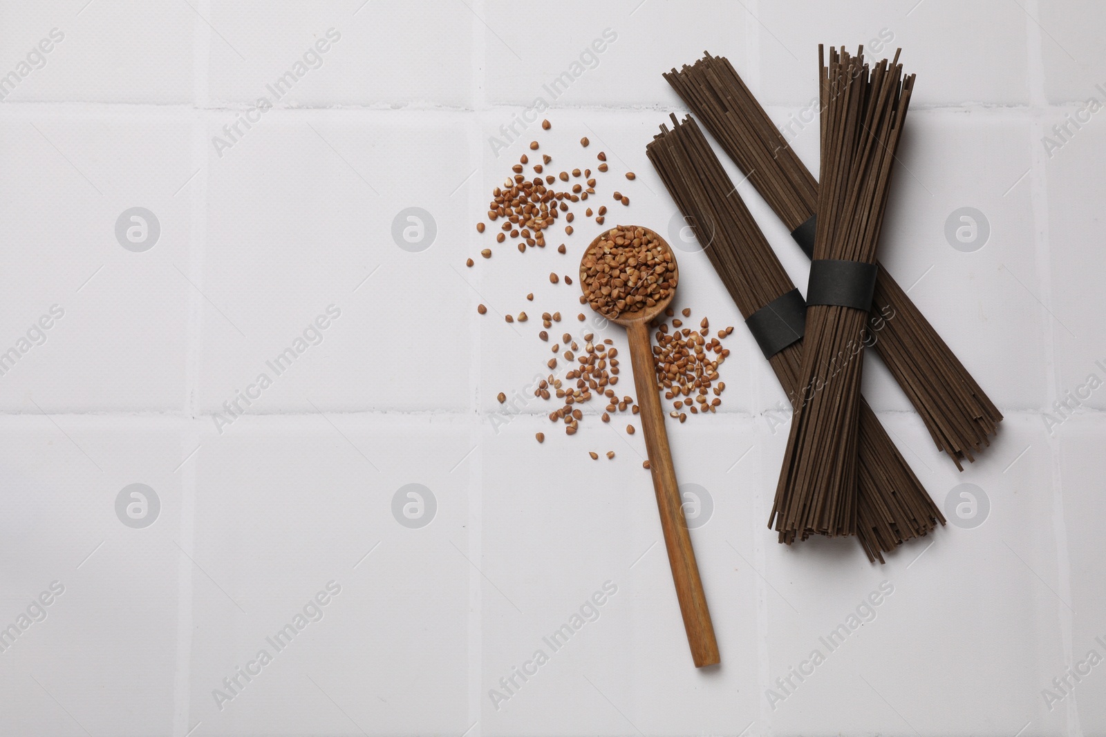 Photo of Uncooked buckwheat noodles (soba), grains and spoon on white tiled table, flat lay. Space for text
