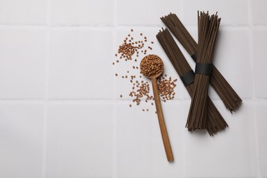 Uncooked buckwheat noodles (soba), grains and spoon on white tiled table, flat lay. Space for text