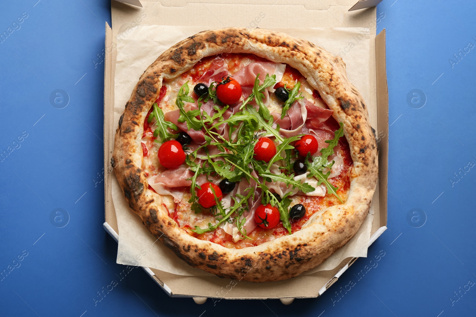 Photo of Tasty pizza with meat and arugula in cardboard box on blue background, top view