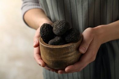 Woman holding wooden bowl of black truffles in hands on beige background, closeup