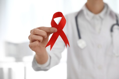 Photo of Doctor holding red ribbon on blurred background, closeup. AIDS awareness month