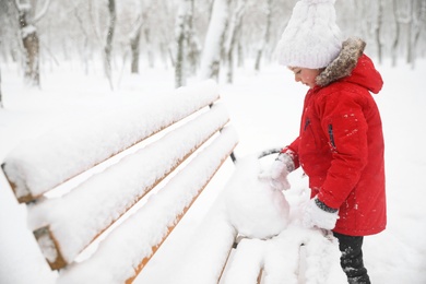 Photo of Cute little girl rolling snowball on bench in winter, space for text