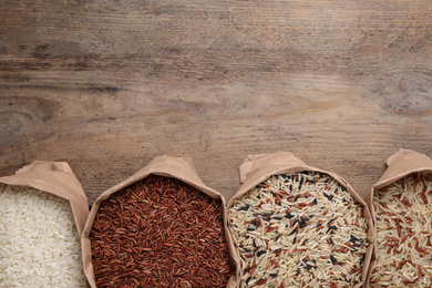 Photo of Different types of brown and polished rice in paper bags on wooden table, flat lay. Space for text