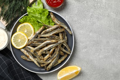 Delicious fried anchovies with lemon, lettuce leaves and sauces served on light grey table, flat lay. Space for text