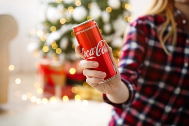 Photo of MYKOLAIV, UKRAINE - January 01, 2021: Woman with can of Coca-Cola against blurred Christmas tree indoors, closeup