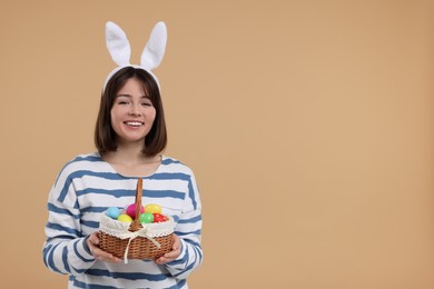 Easter celebration. Happy woman with bunny ears and wicker basket full of painted eggs on beige background, space for text