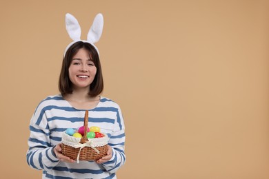 Photo of Easter celebration. Happy woman with bunny ears and wicker basket full of painted eggs on beige background, space for text