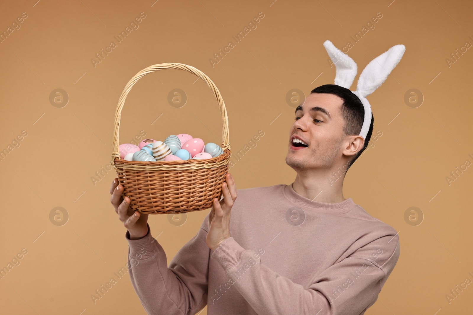 Photo of Easter celebration. Handsome young man with bunny ears holding basket of painted eggs on beige background