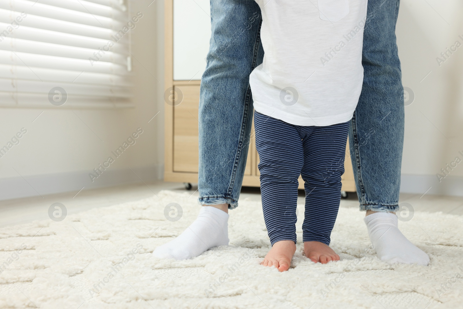Photo of Mother supporting her son while he learning to walk on carpet indoors, closeup. Space for text