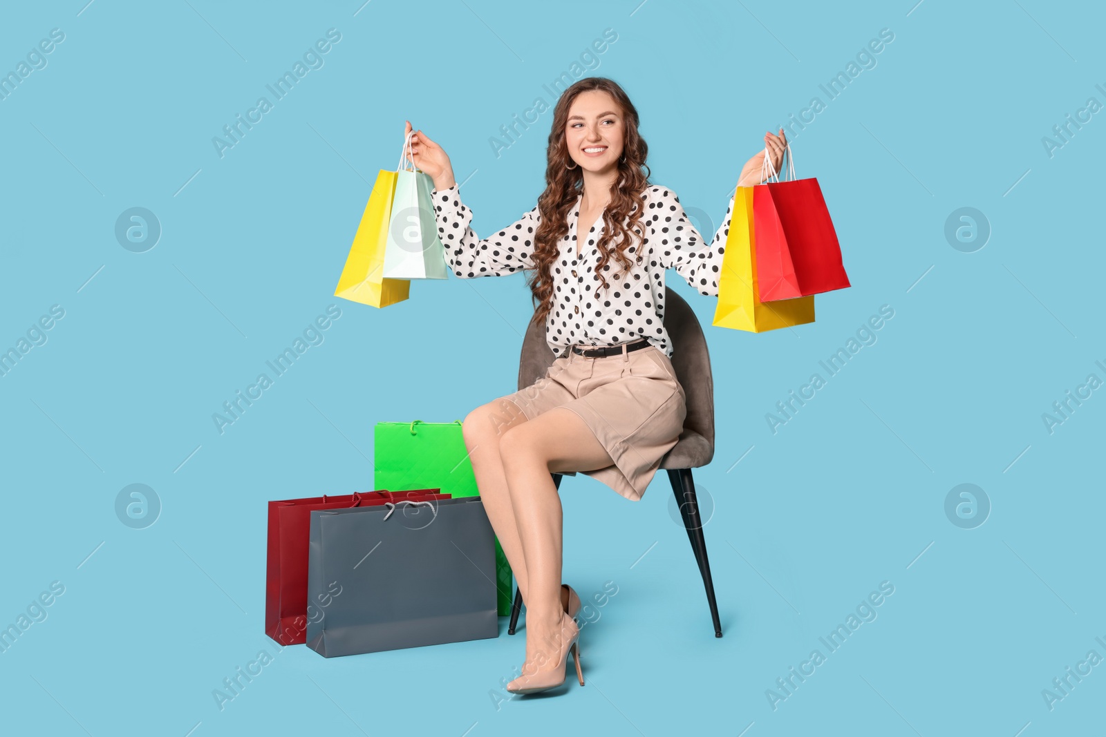 Photo of Happy woman holding colorful shopping bags on chair against light blue background