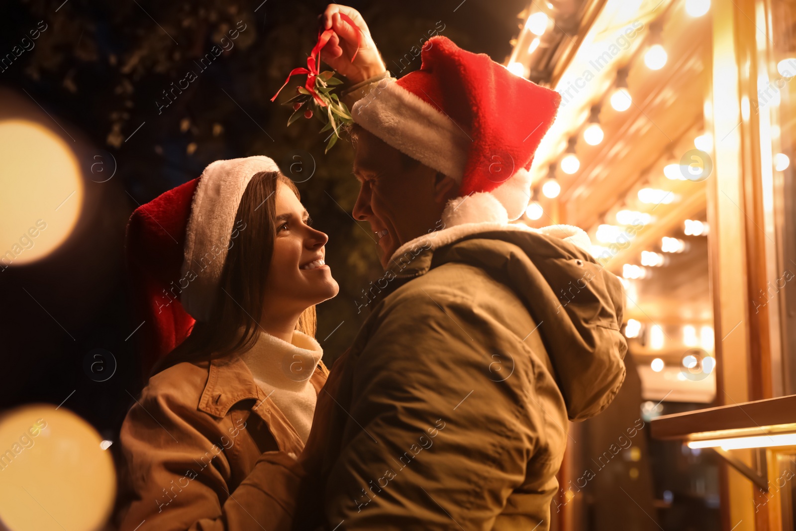 Photo of Happy couple in Santa hats standing under mistletoe bunch outdoors, bokeh effect