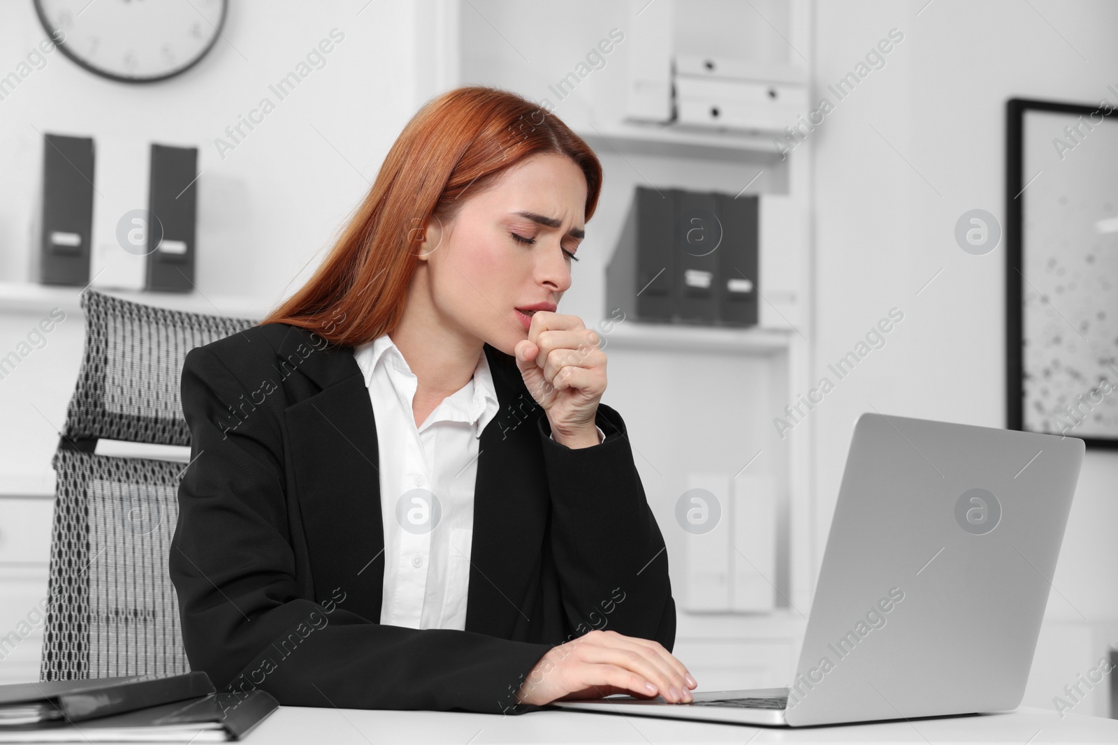 Photo of Woman coughing while using laptop at table in office. Cold symptoms