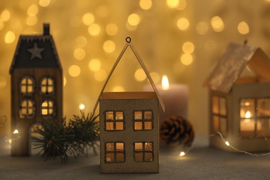 House shaped holders with burning candles on grey table against blurred Christmas lights