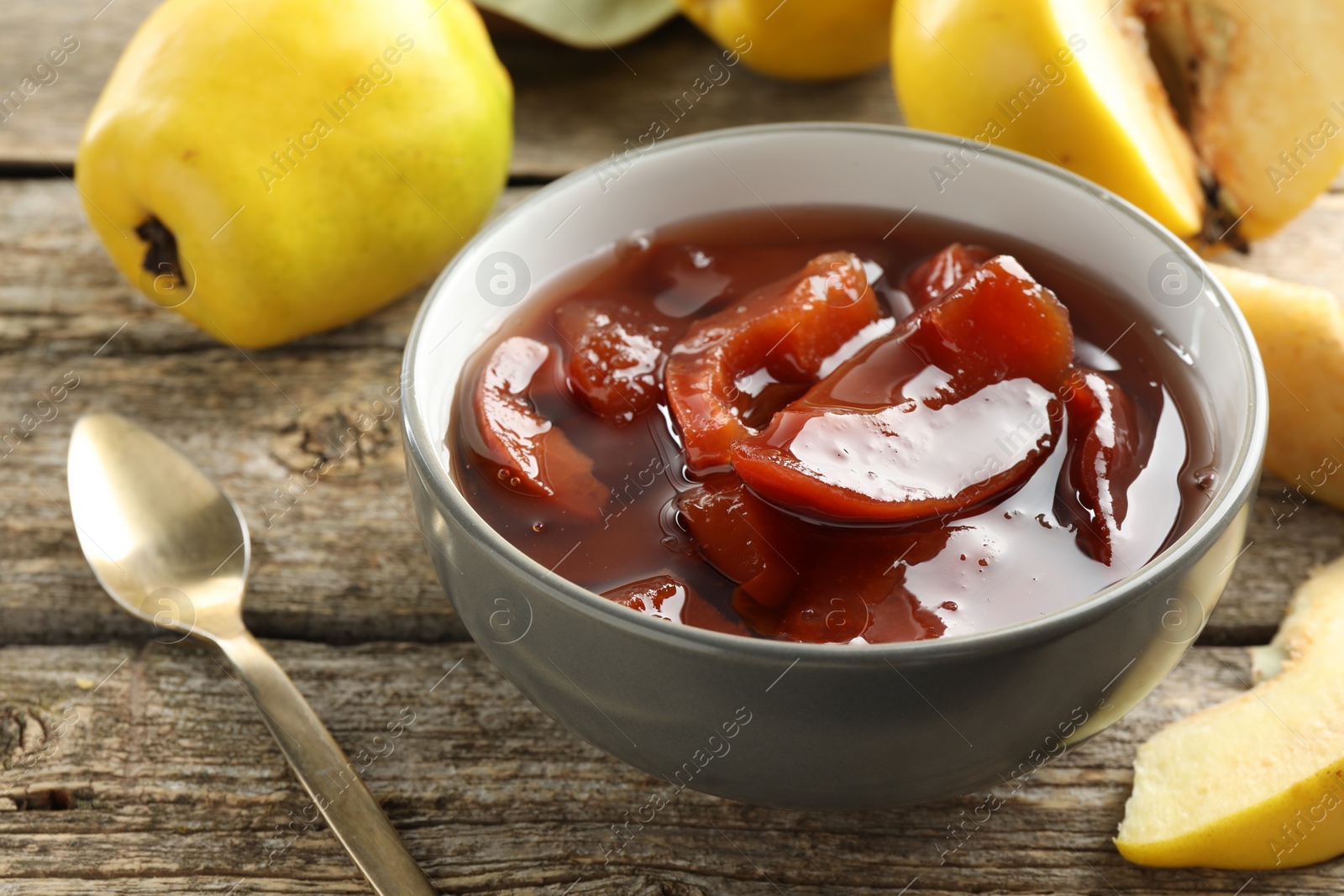 Photo of Tasty homemade quince jam in bowl, spoon and fruits on wooden table, closeup