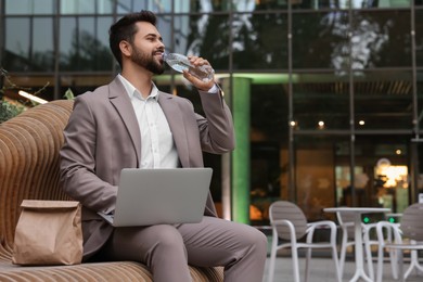 Photo of Young businessman with laptop drinking water from bottle while lunch on bench outdoors