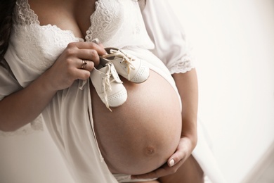 Young pregnant woman in lace nightgown holding baby shoes on light background, closeup. Space for text