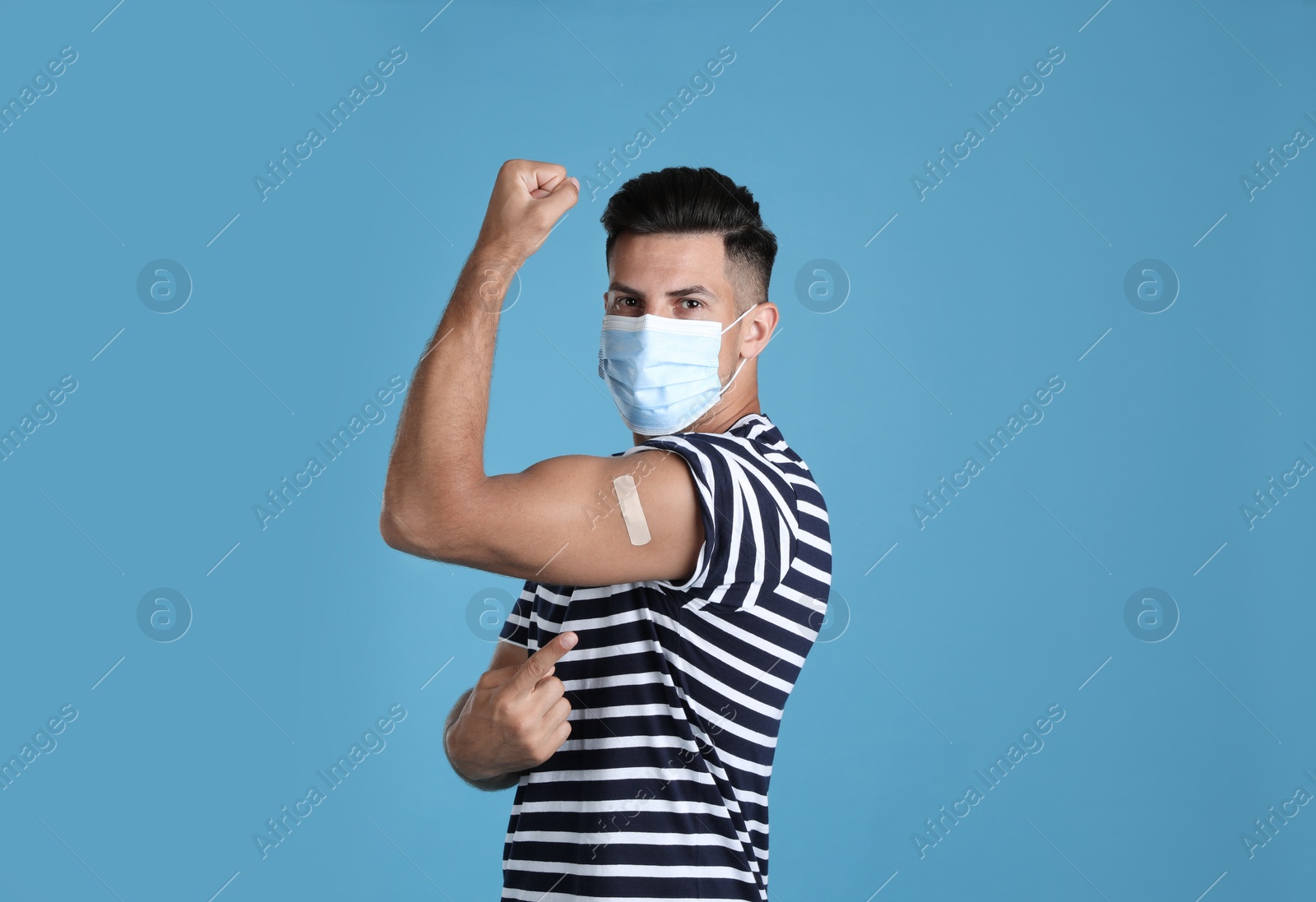 Photo of Vaccinated man with protective mask showing medical plaster on his arm against light blue background