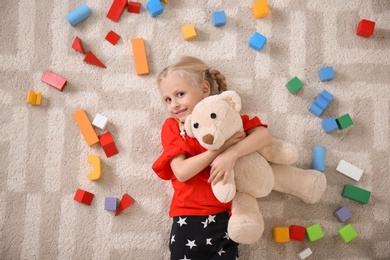 Cute little girl lying on cozy carpet with cubes and teddy bear at home
