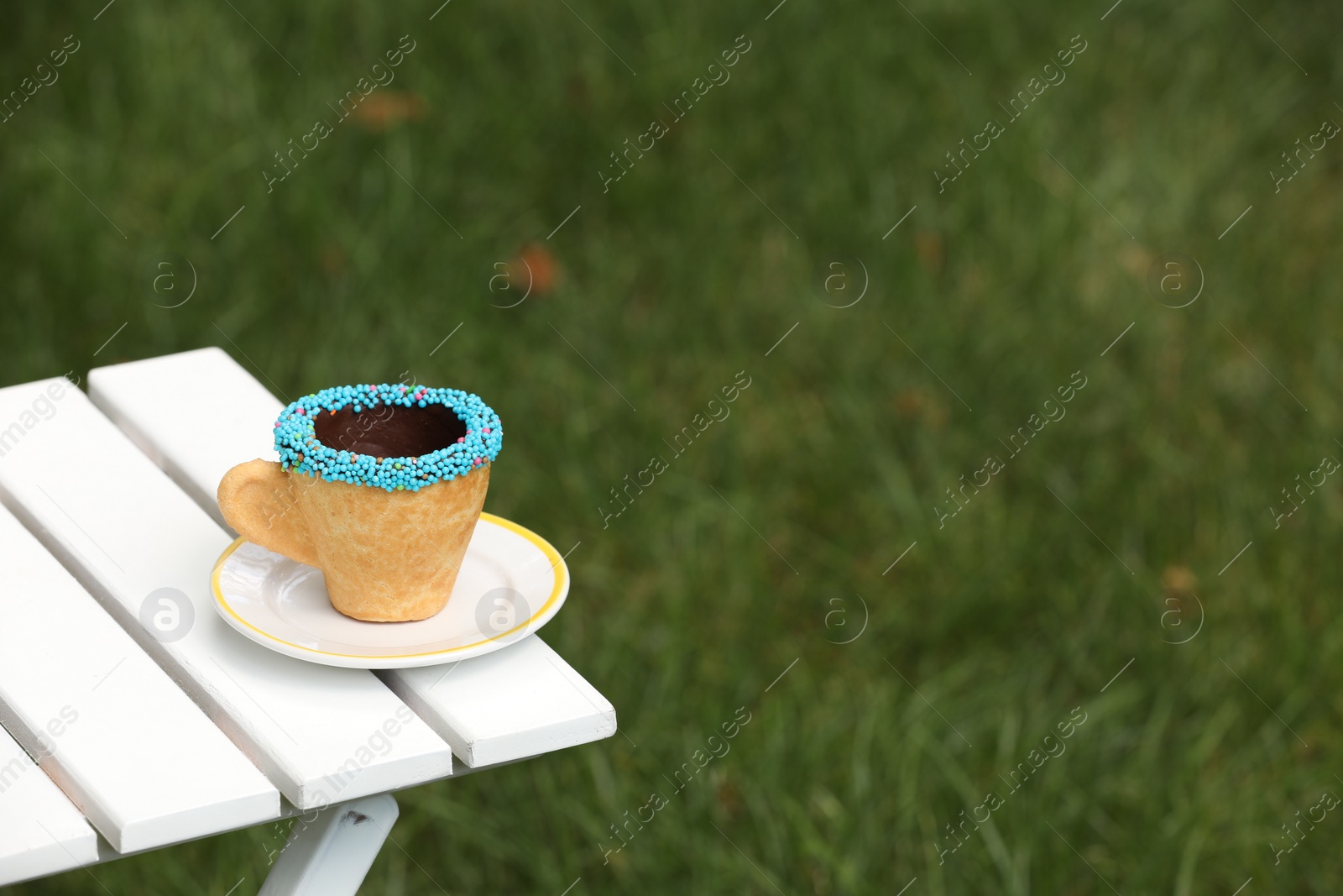 Photo of Delicious edible biscuit coffee cup decorated with sprinkles on white wooden table outdoors, space for text