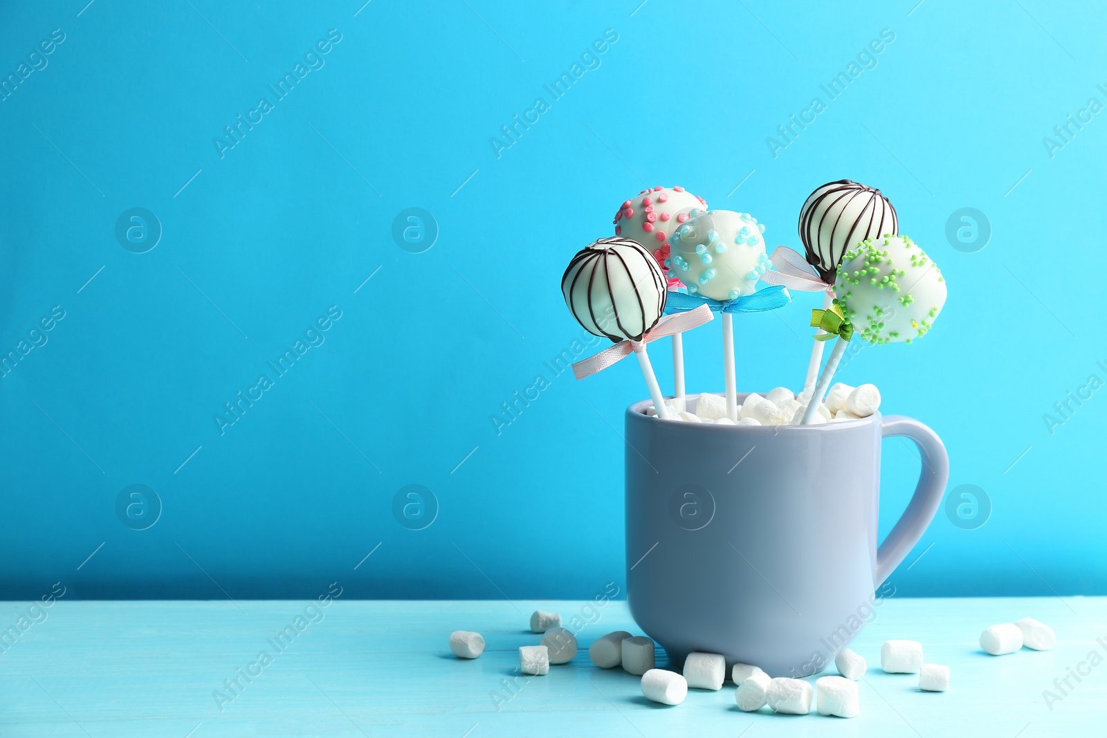 Photo of Tasty cake pops in cup with marshmallow on wooden table against light blue background. Space for text