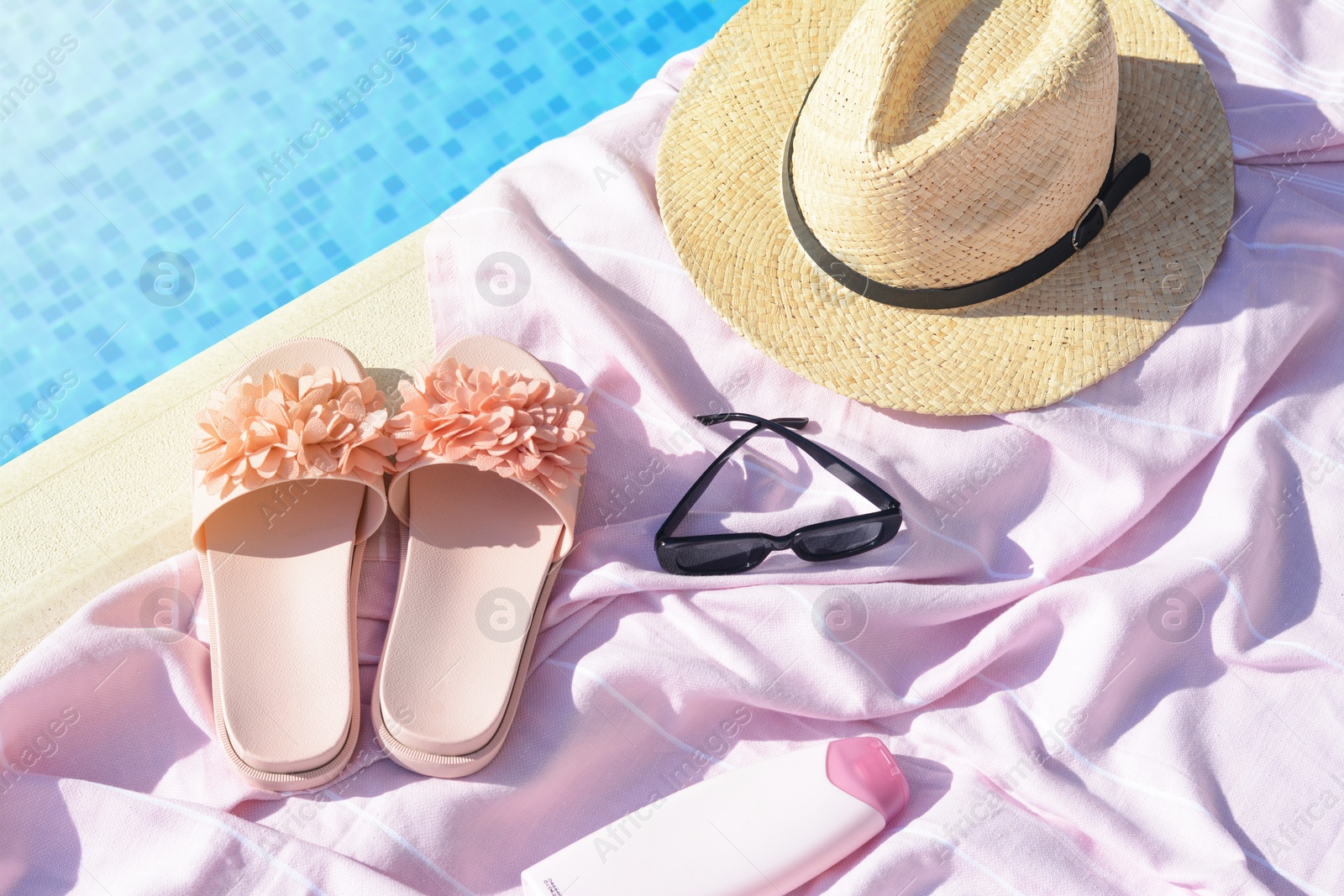 Photo of Pink blanket with slippers, hat, sunglasses and sunscreen near outdoor swimming pool on sunny day