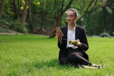 Lunch time. Happy businesswoman with container of salad using smartphone on green grass in park, space for text