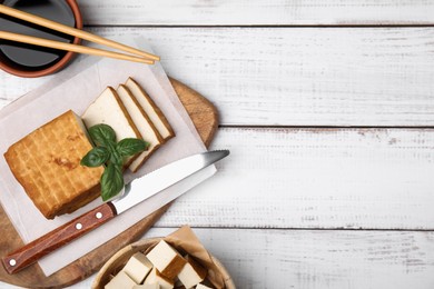 Photo of Board with smoked tofu, knife, basil and soy sauce on white wooden table, flat lay. Space for text