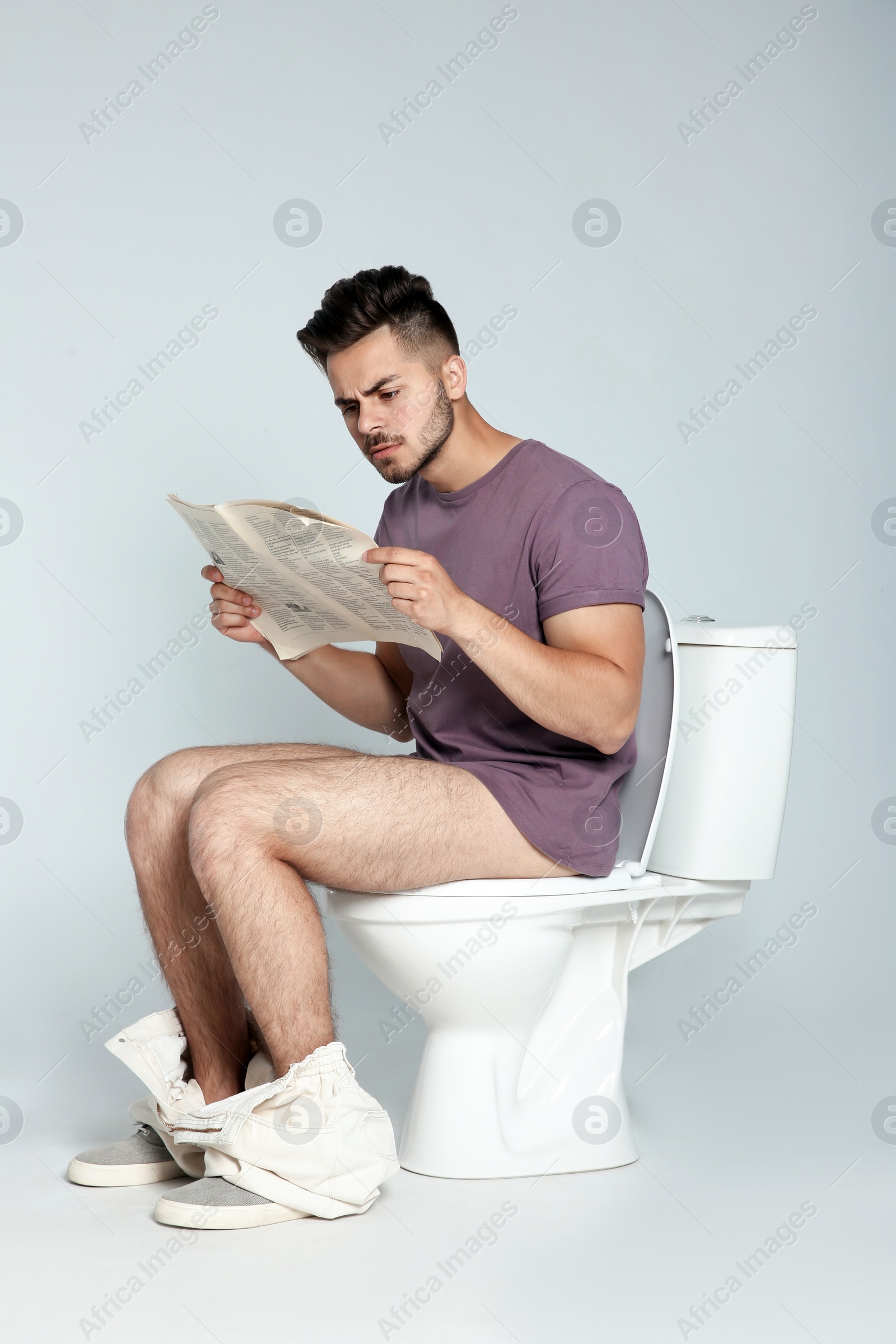 Photo of Young man reading newspaper while sitting on toilet bowl against gray background