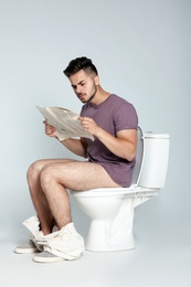 Photo of Young man reading newspaper while sitting on toilet bowl against gray background