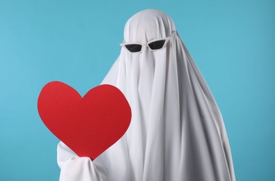 Photo of Cute ghost. Person covered with white sheet in sunglasses holding red heart on light blue background