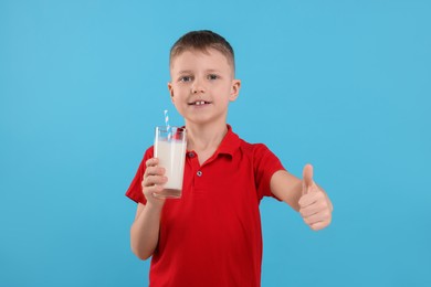 Photo of Cute boy with glass of fresh milk showing thumb up on light blue background