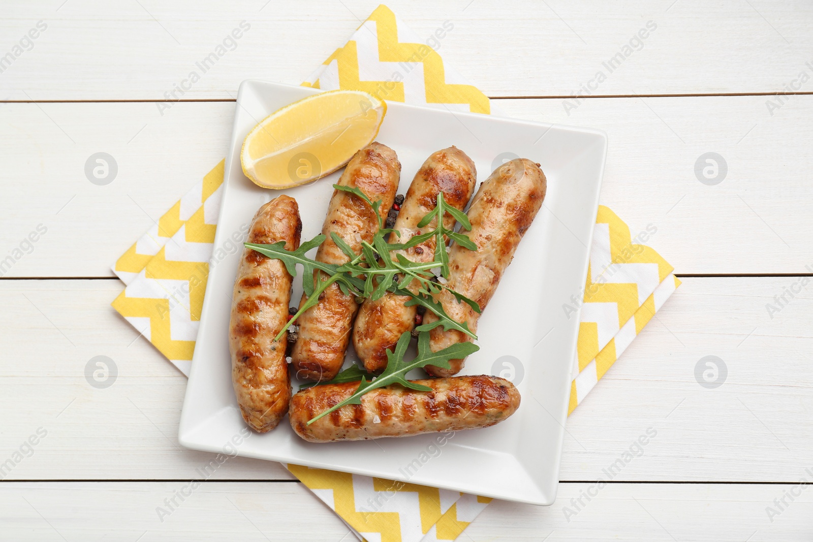 Photo of Tasty grilled sausages served with lemon and arugula on white wooden table, top view